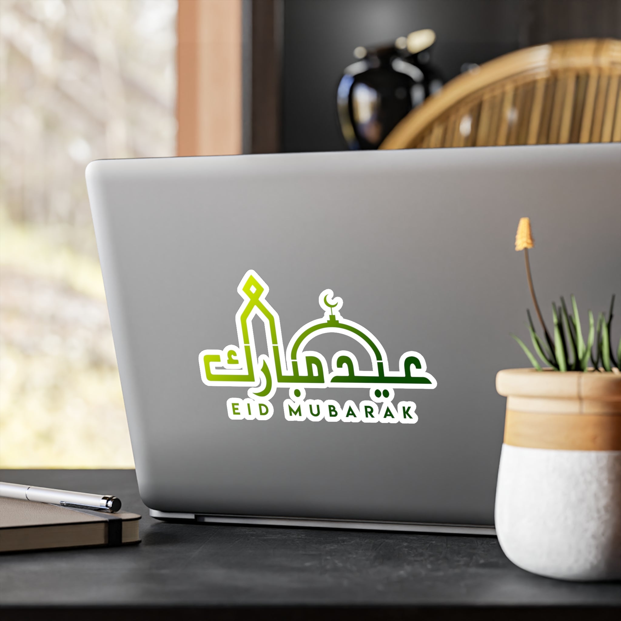 A laptop with a festive "Eid Mubarak" transparent sticker from Medina Roots on the lid, placed on a desk by a window.