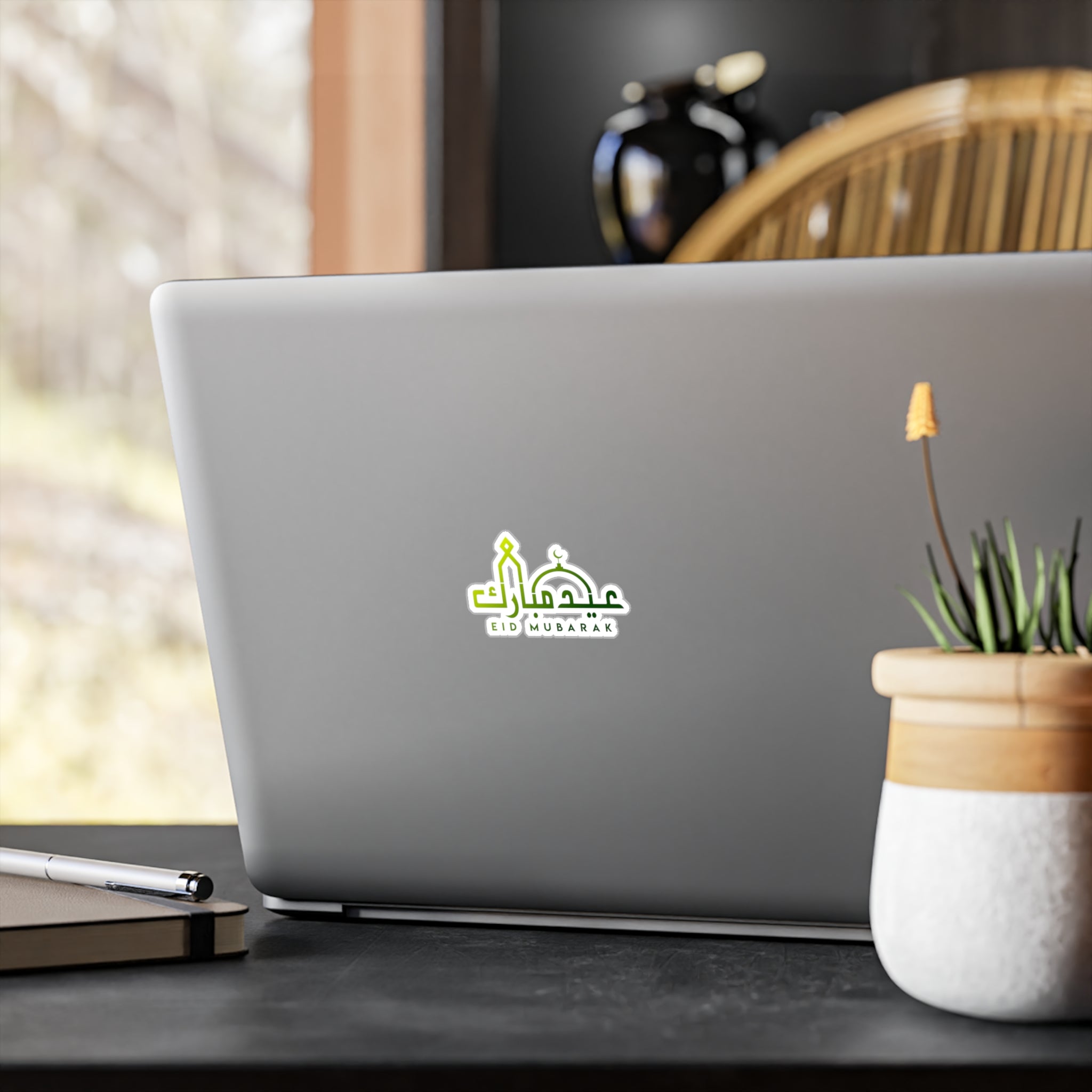 A laptop on a wooden desk with a Medina Roots Eid Mubarak Transparent Outdoor Sticker on its lid, next to a potted plant and a smartphone.