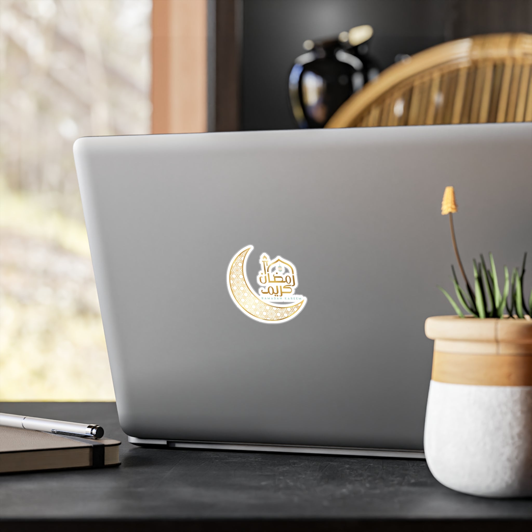 A laptop with a Ramadan Kareem Transparent Outdoor Sticker from Medina Roots, symbolizing Ramadan, on its lid, placed on a table near a window with outdoor views.