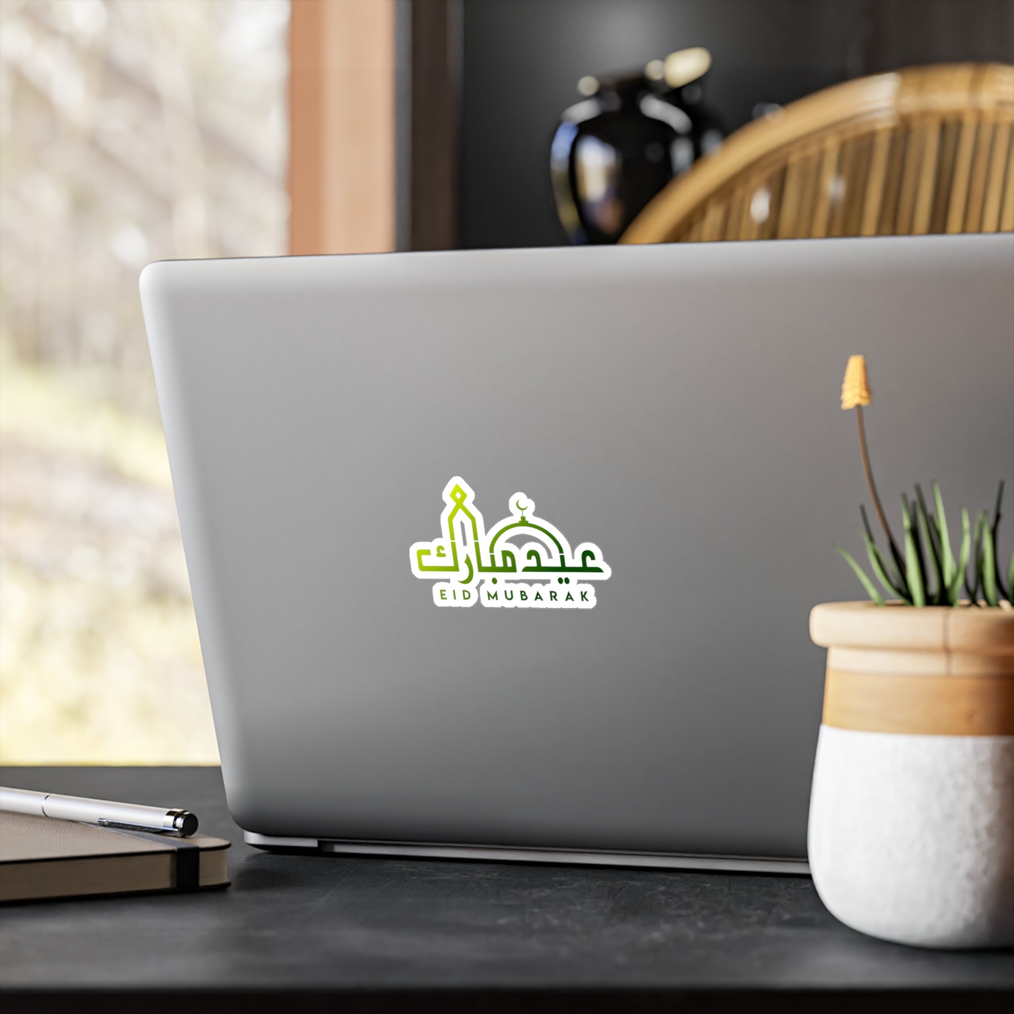 A laptop with a 'Medina Roots Eid Mubarak Transparent Outdoor Sticker' on its lid, placed on a desk near a window.