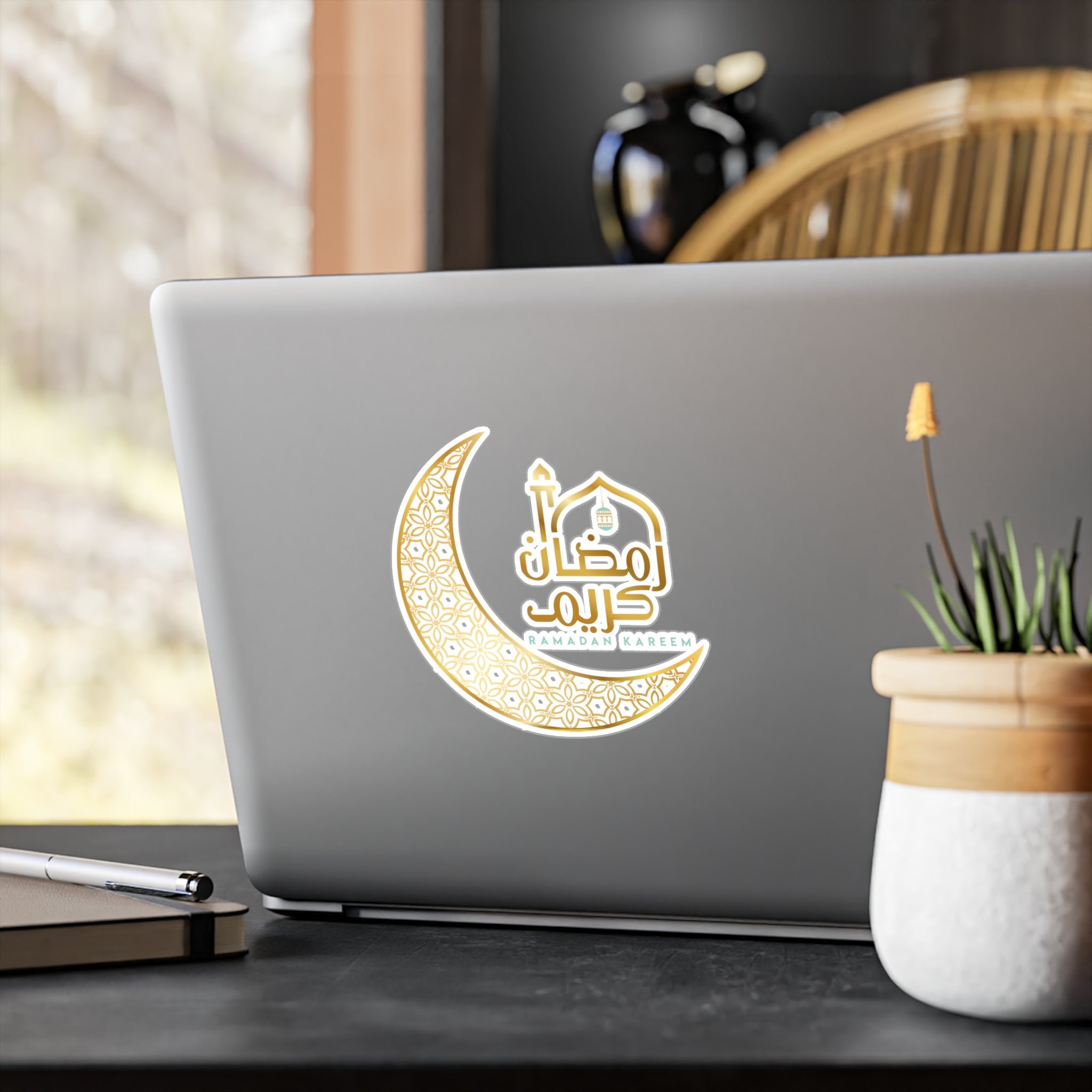 Laptop with a Medina Roots Ramadan Kareem Transparent Outdoor Sticker celebrating Ramadan on its lid, placed on a wooden table beside a potted plant.