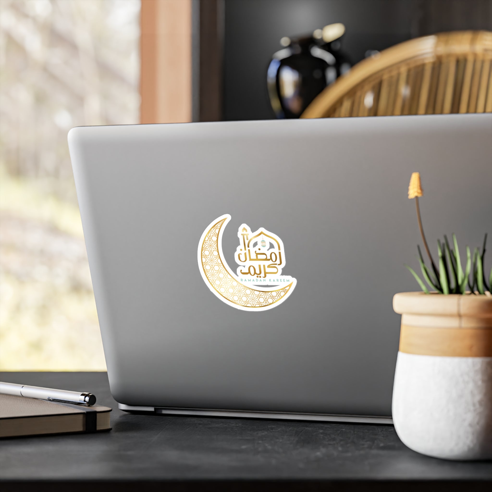 A laptop with a Medina Roots Ramadan Kareem Transparent Outdoor Sticker featuring arabic calligraphy moon on its lid, placed on a desk next to a potted plant.