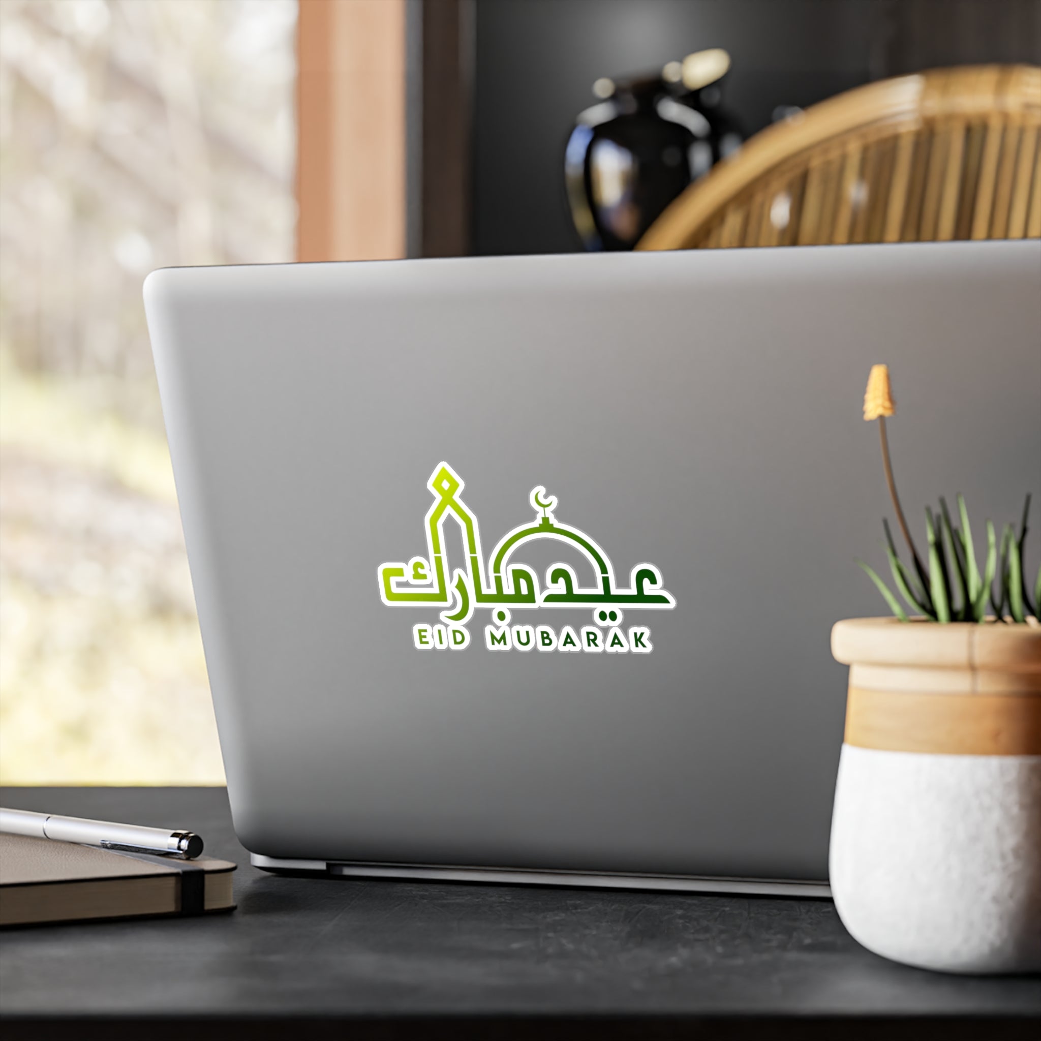 Laptop with a 'Medina Roots Eid Mubarak Transparent Outdoor Sticker' on the lid, placed on a wooden desk next to a potted plant.