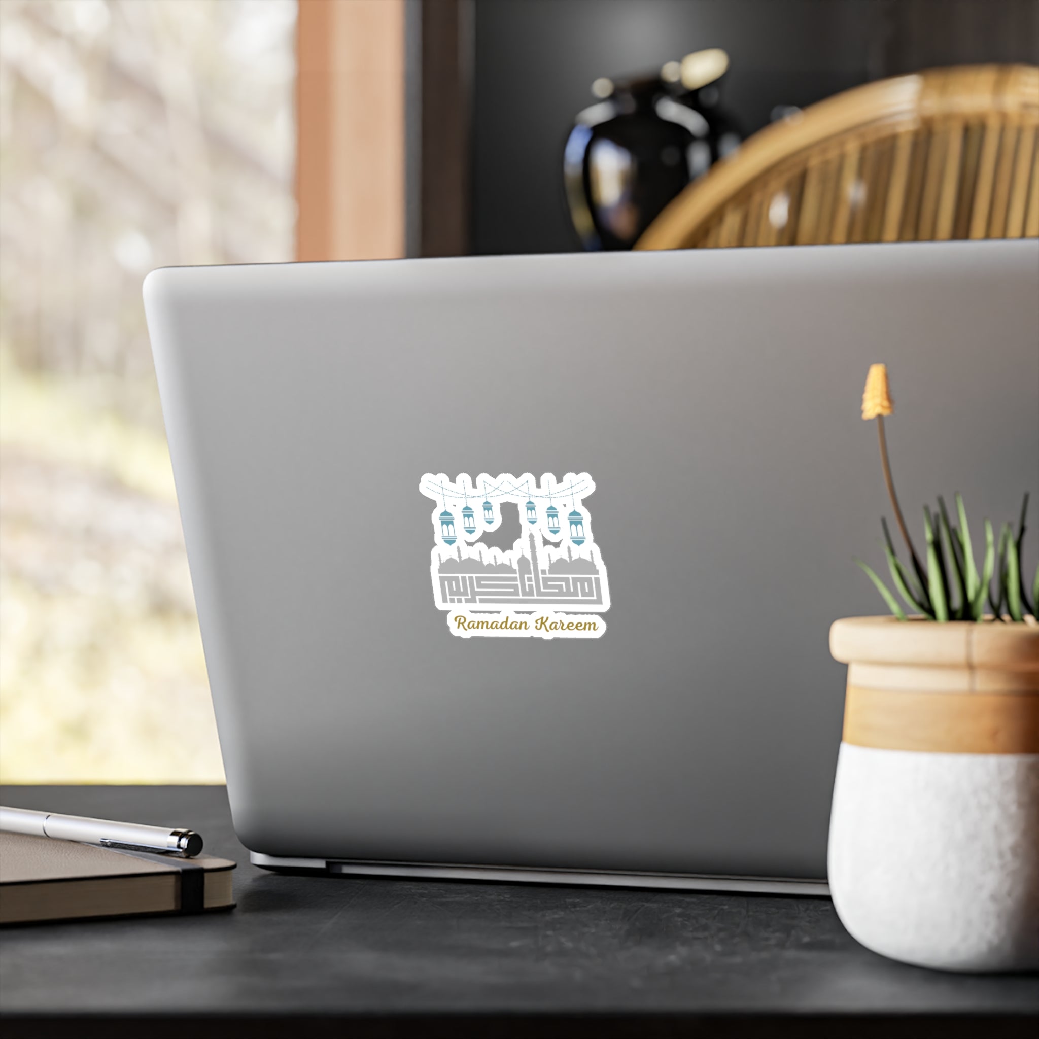 A laptop with a Medina Roots Ramadan Kareem Kiss-Cut Vinyl Decal on its lid, placed on a wooden table near a window with a plant beside it.